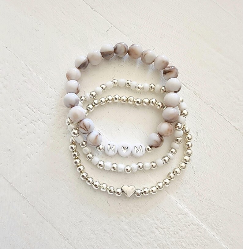 May include: Set of four bracelets with white and brown marbled beads, silver beads, and silver letter beads spelling "MOM" with a heart between the letters. The bottom bracelet has a silver heart charm.