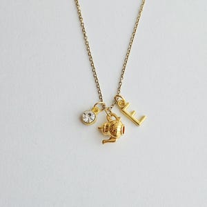 May include: A gold-colored necklace with three charms: a clear crystal, a small teapot, and the letter "E". The necklace hangs on a delicate gold chain against a white background.