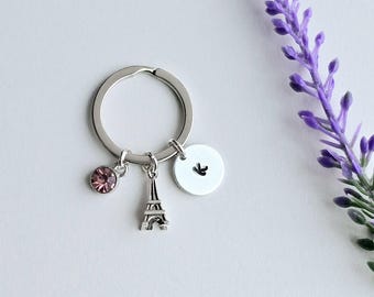May include: A silver keychain with a pink crystal charm, a miniature Eiffel Tower charm, and a silver disc charm with a small bird design.