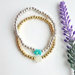 May include: Two beaded bracelets with silver and gold beads. One bracelet has a turquoise elephant charm and the other has a white elephant charm.
