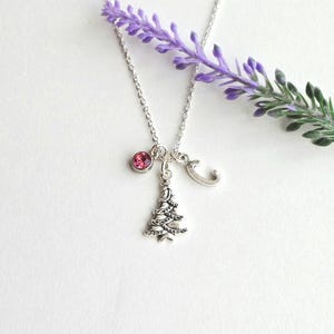 May include: A silver necklace with a Christmas tree charm, a pink gemstone, and a letter "C" charm. The necklace is displayed on a white surface with a sprig of purple flowers.