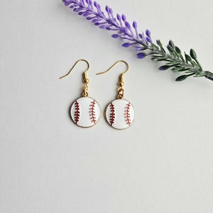 May include: A pair of baseball-themed earrings. Each earring features a white, circular design with red stitching, resembling a baseball. The earrings have gold-colored hooks. A sprig of lavender and greenery is in the background.