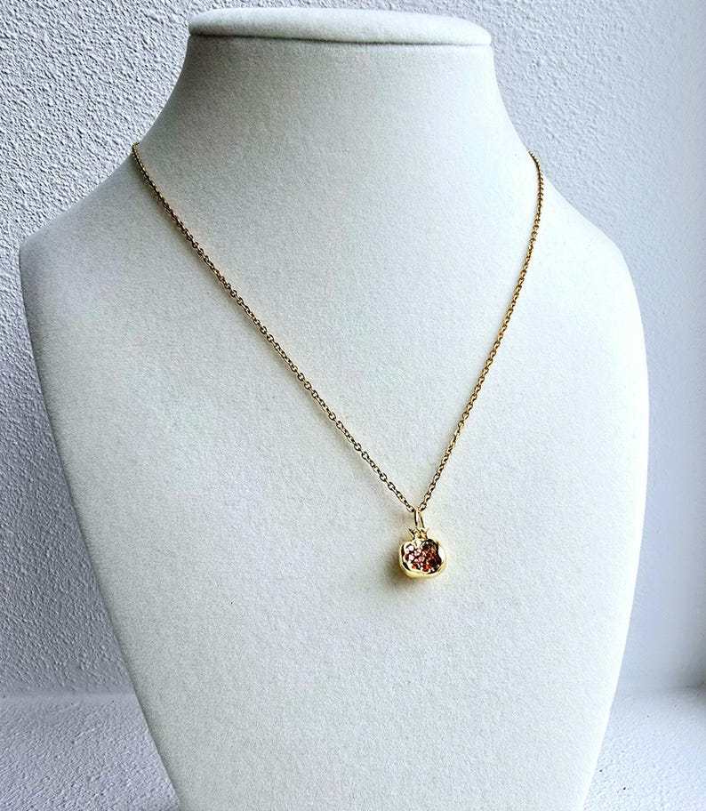 K&ouml;nnte beinhalten: Eine goldene Kettenkette mit einem kleinen, strukturierten, rot-goldenen Anh&auml;nger in Form eines Apfels.