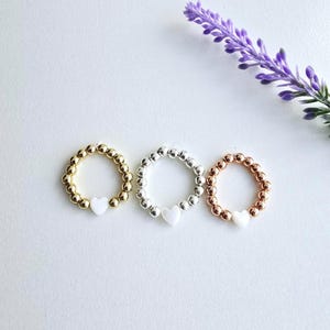 May include: Three beaded rings with a white heart charm. The rings are gold, silver, and rose gold.