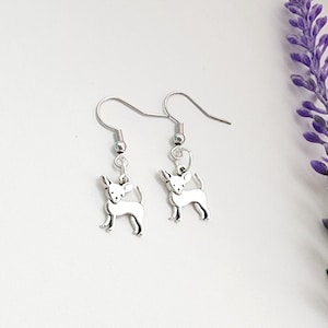 May include: A pair of silver-colored Chihuahua dog earrings. Each earring features a small, detailed charm of a Chihuahua dog suspended from a fish hook ear wire. The earrings are displayed on a white surface with a purple flower in the background.