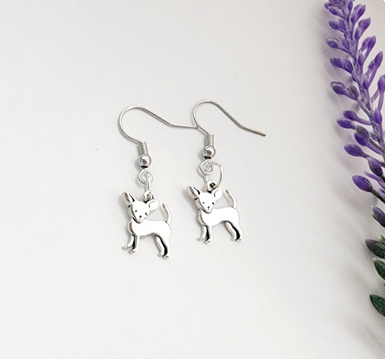 May include: A pair of silver-colored Chihuahua dog earrings. Each earring features a small, detailed charm of a Chihuahua dog suspended from a fish hook ear wire. The earrings are displayed on a white surface with a purple flower in the background.