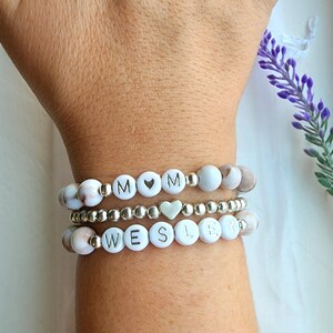 May include: Three white beaded bracelets with silver beads and silver lettering. The top bracelet spells "MOM" with a silver heart bead between the letters. The bottom bracelet spells "WESLEY".