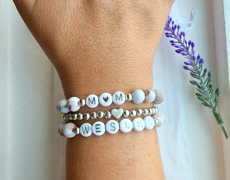 May include: Three white beaded bracelets with silver beads and silver lettering. The top bracelet spells "MOM" with a silver heart bead between the letters. The bottom bracelet spells "WESLEY".