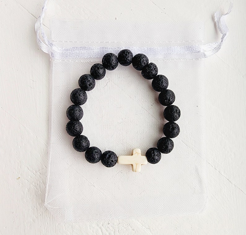 May include: A black lava rock beaded bracelet with a white cross charm. The bracelet is in a white organza bag.