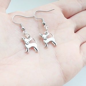 May include: Silver-colored Chihuahua dog earrings. Each earring features a small, detailed Chihuahua charm hanging from a silver-tone fish hook. The charms are in a standing pose with their tails curled.