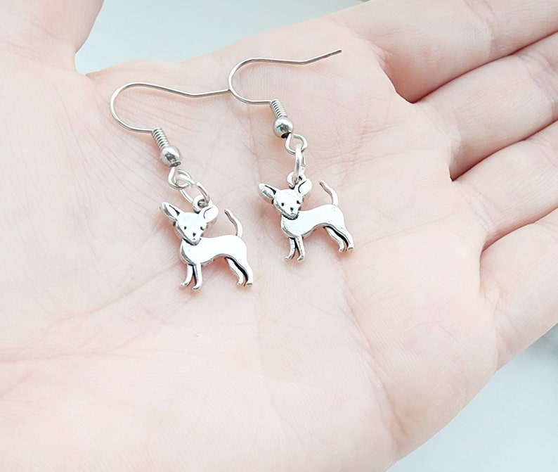 May include: Silver-colored Chihuahua dog earrings. Each earring features a small, detailed Chihuahua charm hanging from a silver-tone fish hook. The charms are in a standing pose with their tails curled.