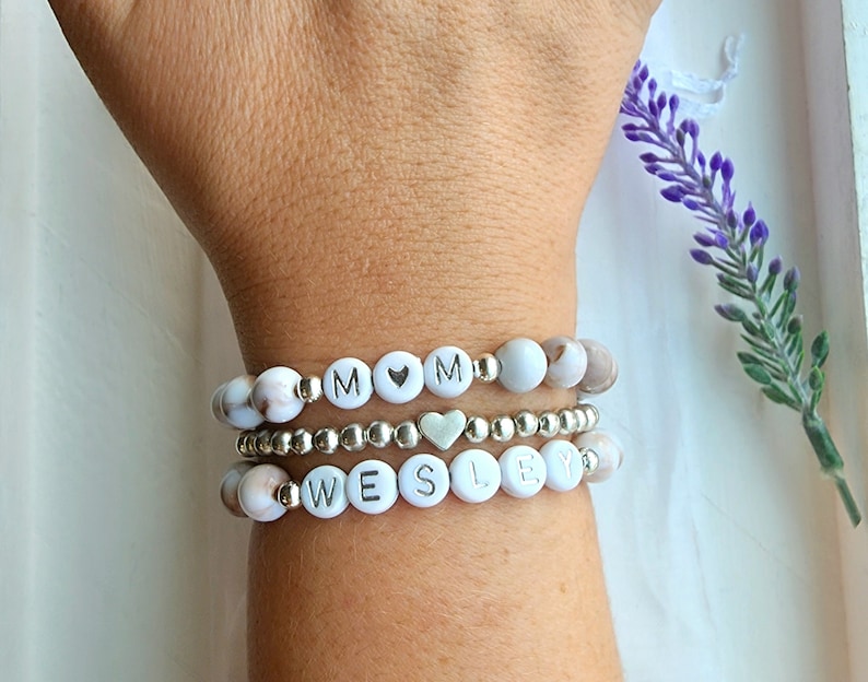 May include: Three bracelets stacked on a wrist. The top bracelet has white beads with the letters "MOM" and a silver heart charm. The middle bracelet is a silver chain. The bottom bracelet has white beads with the letters "WESLEY".