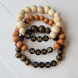 May include: Three stacked bracelets made of wooden beads in cream, brown, and green colors. Each bracelet has the word "MAMA" spelled out in black beads with gold lettering.