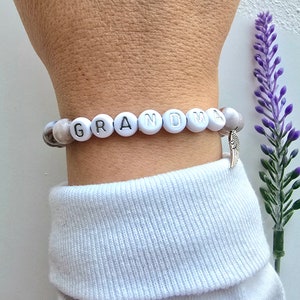 Puede incluir: Un brazalete de cuentas blancas con la palabra "GRANDMA" escrita en letras plateadas. El brazalete tiene un peque&ntilde;o dije de ala de &aacute;ngel plateado.