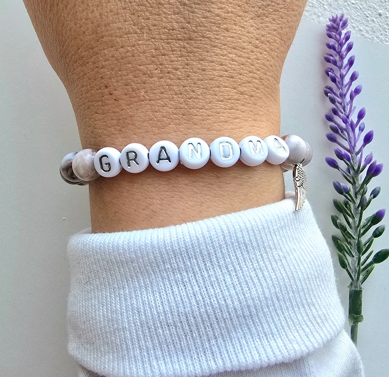 Puede incluir: Un brazalete de cuentas blancas con la palabra "GRANDMA" escrita en letras plateadas. El brazalete tiene un peque&ntilde;o dije de ala de &aacute;ngel plateado.