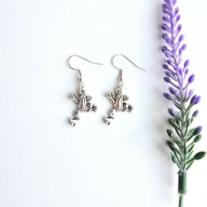 May include: Silver frog earrings with a dangling design. The earrings are on a white background with a sprig of purple flowers.