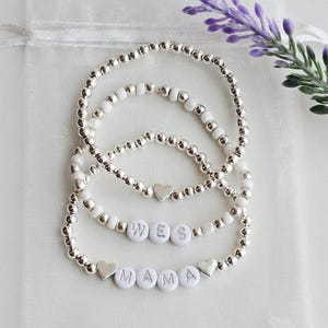 May include: A set of three stretch bracelets featuring metallic silver-tone beads and white beads. One bracelet includes a silver-tone heart charm, another features white letter beads spelling WES, and the third displays MAMA with heart charms.