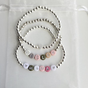 May include: Three stretch bracelets made of small metallic silver beads. One bracelet features a silver heart charm. Another bracelet includes round beads spelling the name WESLEY in white, gray, pink, and olive green. The third spells MAMA with hearts.