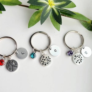 May include: Three silver keychains with charms. Each keychain has a silver circle with a letter, a crystal, and a silver charm with a constellation design. The constellations are Cancer, Gemini, and Aquarius. The crystals are red, blue, and purple.