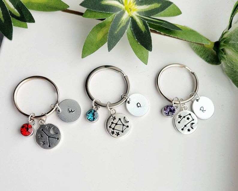 May include: Three silver keychains with charms. Each keychain has a silver circle with a letter, a crystal, and a silver charm with a constellation design. The constellations are Cancer, Gemini, and Aquarius. The crystals are red, blue, and purple.