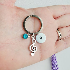 May include: Silver keychain with a treble clef charm, a teal-colored crystal, and a small, round, silver charm with the letter "G". The keychain is held in a hand.