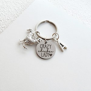 May include: Silver-toned keychain featuring a chicken charm, a circular pendant with the text "Crazy Chicken Lady" and a heart, and a letter "A" charm. The keychain is on a silver ring, and the background is white.