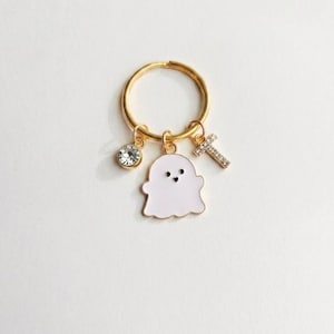May include: A gold-toned metal keyring featuring three charms: a white enamel ghost with black eyes and a heart-shaped mouth, a small circular clear rhinestone charm, and a gold-toned letter T charm encrusted with small clear rhinestones.