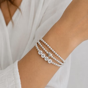 May include: A stack of three silver-toned beaded bracelets. One bracelet features white circular beads spelling out the word MAMA in black letters, while another bracelet includes a small silver-toned heart charm among the metallic beads.