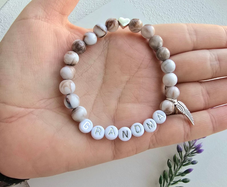 Puede incluir: Pulsera de cuentas blancas con cuentas marmoleadas marrones, un dije de coraz&oacute;n plateado y un dije de ala de &aacute;ngel plateado. La pulsera tiene la palabra "GRANDMA" escrita en letras plateadas sobre cuentas blancas.
