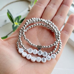May include: Two stacked stretch bracelets made of small, shiny silver-toned metallic beads. One bracelet features a flat silver heart charm, while the other includes white round beads with black letters spelling out the word UNIQUELY BIRR.