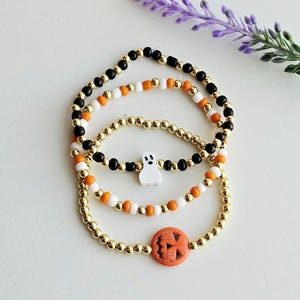 May include: Three Halloween-themed beaded bracelets. One bracelet features a white ghost charm, another has an orange pumpkin charm. The bracelets are gold, black, white, and orange. A sprig of purple flowers is in the background.