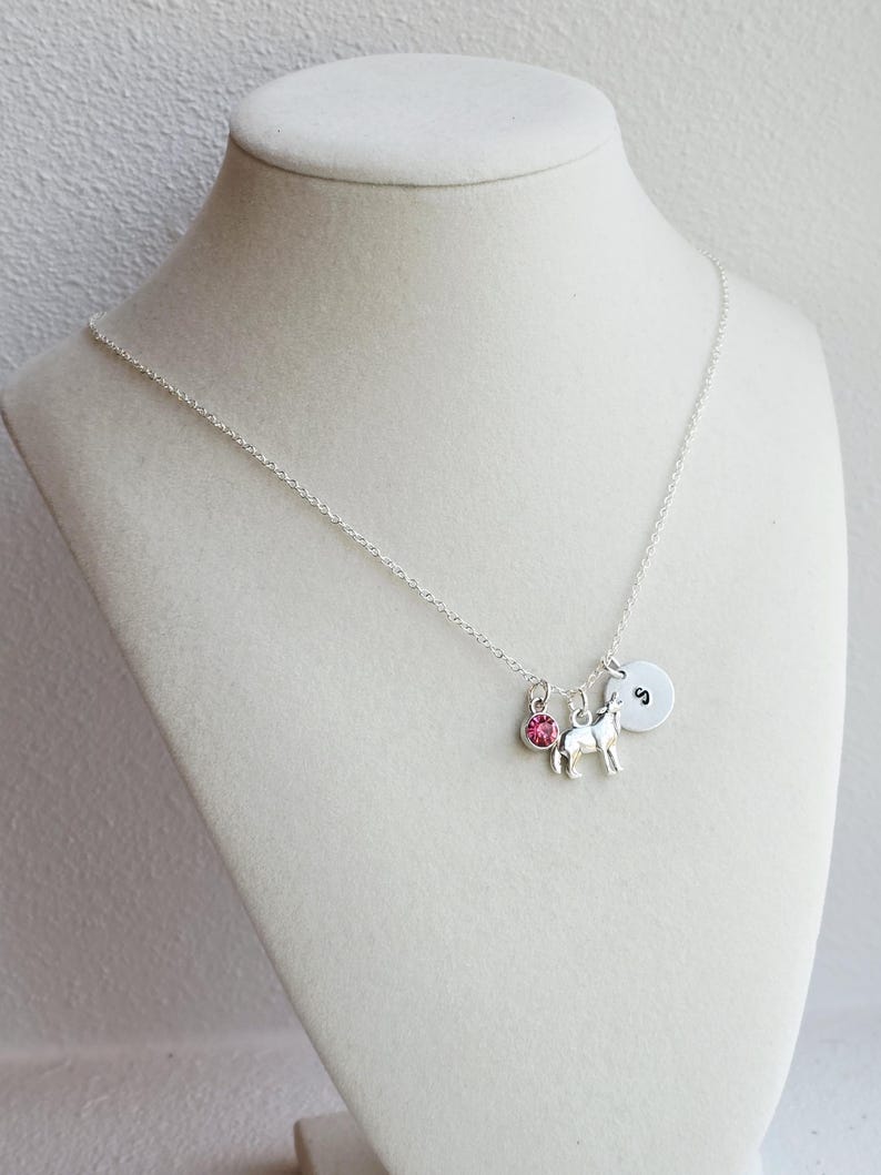 Peut inclure: Un collier argent&eacute; avec trois breloques : une pierre rose, un petit cheval et un charm rond avec la lettre "S". Le collier est pr&eacute;sent&eacute; sur un mannequin blanc.