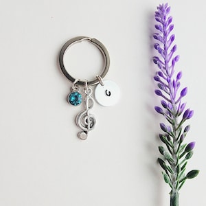 May include: Silver keychain with a treble clef charm, a teal-colored crystal, and a small round tag with the letter "G". A sprig of purple lavender is on the right side of the image. The keychain is on a white background.