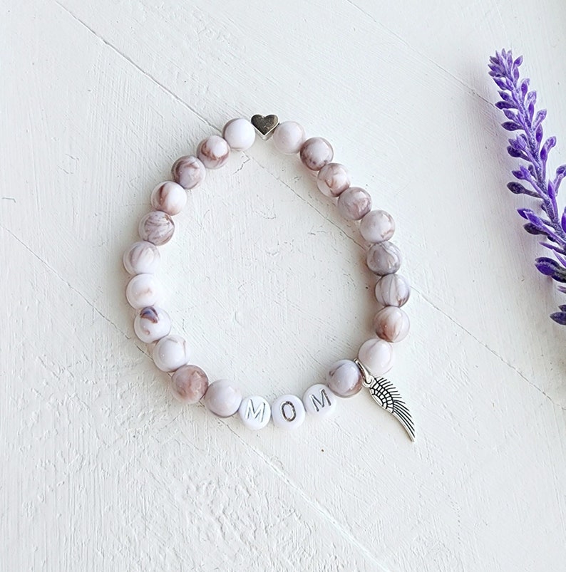 Puede incluir: Pulsera de cuentas de m&aacute;rmol blanco y marr&oacute;n con un dije de coraz&oacute;n plateado y un dije de ala de &aacute;ngel plateado. La pulsera tambi&eacute;n tiene cuentas blancas con las letras "MOM" deletreadas.