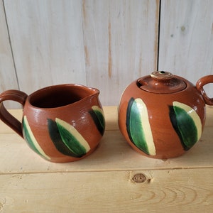 May include: A brown ceramic creamer and sugar set with green and yellow leaf designs.