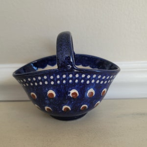 May include: A blue ceramic bowl with a handle, decorated with white dots and brown and white circles. The bowl is shaped like a basket.