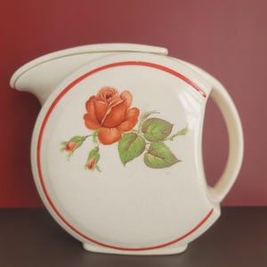 May include: Cream-colored ceramic pitcher with a red rose design. The pitcher has a curved handle and a red trim. The rose is orange with green leaves and buds. The pitcher is a vintage style.