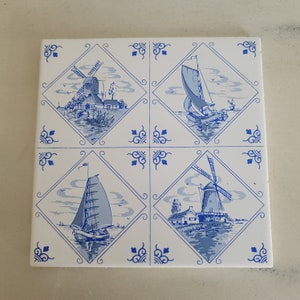 May include: A white ceramic tile with a blue and white design featuring four Dutch scenes. Each scene is set within a diamond shape and features a windmill and a sailboat. The tile has a decorative border with blue swirls.