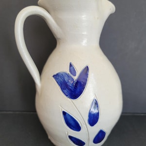 May include: A cream-colored ceramic pitcher with a curved handle and a spout. The pitcher is decorated with a blue floral design. The pitcher is a decorative item.