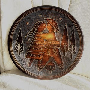 May include: Round wooden plate with a carved design. The plate features two figures embracing, surrounded by trees and stars. The figures and trees are a lighter brown against a darker brown background. The plate has a gold-colored border.