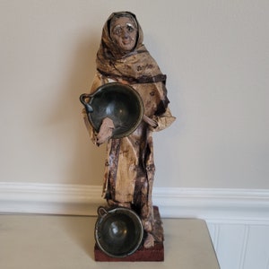 May include: A handmade figurine of a woman wearing a brown and white robe, holding two small, round, dark green bowls. The figurine is standing on a wooden base.