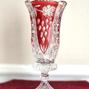 May include: A clear crystal vase with a red interior. The vase has a flared top with a floral design and a series of teardrop shapes. The vase is approximately 10 inches tall and sits on a pedestal base.