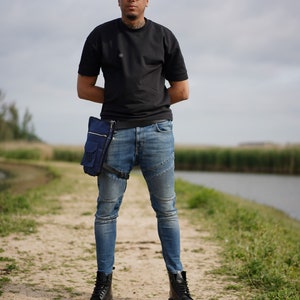 Op de afbeelding: Een persoon draagt een zwart T-shirt, een blauwe jeans en zwarte laarzen. Ze dragen een blauwe heuptas aan hun rechterheup. De persoon staat op een zandpad met groen gras en een waterpartij op de achtergrond.
