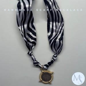 May include: A handmade scarf necklace with a black and white zebra print scarf. The necklace features a gold-colored pendant with a detailed design. The text "HANDMADE SCARF NECKLACE" is printed at the top.