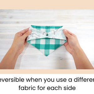 Op de afbeelding: Een omkeerbare groene en witte geruite hondenbandana met een witte stof aan de andere kant met een zwart-witte kattenprint. De bandana is dubbelgevouwen en wordt vastgehouden door een hand.