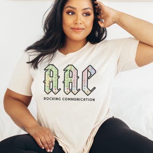 May include: A cream-colored t-shirt with the text "AAC ROCKING COMMUNICATION" in a colorful, stylized font. The graphic features a grid-like design with various symbols and colors. The shirt is a casual, everyday wear item.