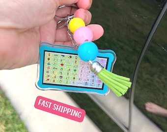 May include: A blue and white keychain with a grid of colorful symbols. The keychain has a green tassel and three beads: yellow, pink, and blue.