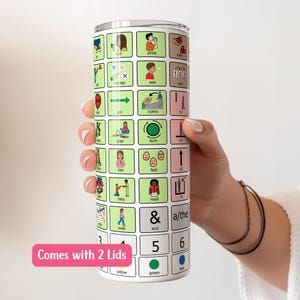 May include: A stainless steel tumbler with a green and white design featuring various images and words. The tumbler has a clear lid and comes with two lids. The text on the tumbler includes "Comes with 2 Lids", "yellow", "green", "blue", "&", "a/the", "and", "5", "6", "walk", "help", "read", "in", "feel", "like", "play", "turn", "on", "go", "come", "to", "see", "choose", "123", "drink", "eat", "mom".