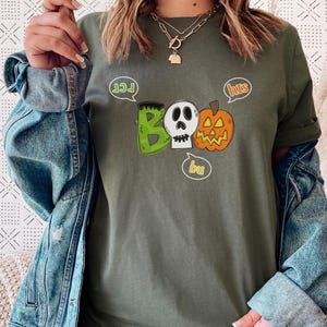 May include: A green, gray, and orange t-shirt with a graphic of a ghost, a pumpkin, and a monster with the words "Boo" and "hits" in speech bubbles.