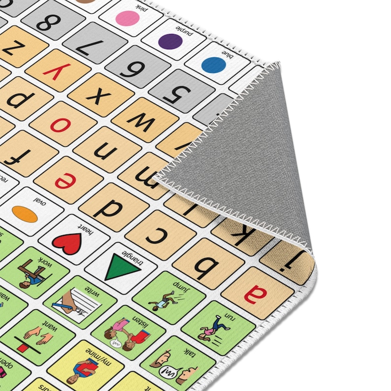 An AAC core vocabulary rug is on white floor.  The edge of the AAC carpet is shown.